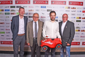 Drei Männer posieren vor einer Sponsorwand und präsentieren ein rotes Spielzeugauto als Fotomotiv bei einem Vereins-Event.