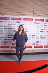 Stehende Frau auf rotem Teppich vor einer Sponsorwand mit Logos; präsentiert sich bei einer Veranstaltung.
