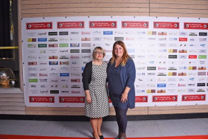 Zwei Personen stehen vor einer Wand mit vielen Vereins-Logos und Sponsorplaketten; Holzrahmen im Hintergrund, für Gruppenfoto.