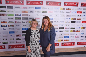 Zwei Frauen posieren lächelnd nebeneinander vor einer Sponsorwand, im Hintergrund vielfarbige Logos; Aufnahme für Event-Teilnahme.