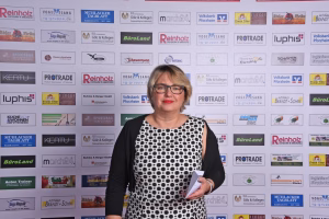 Portrait einer Frau vor einer Wand mit Logos; sie hält Unterlagen und blickt in die Kamera, dokumentiert einen Event/Pressetermin.