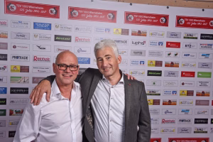 Zwei Männer posieren eng beieinander vor einer Sponsorwand und lächeln in die Kamera, mit Arm auf Schulter.