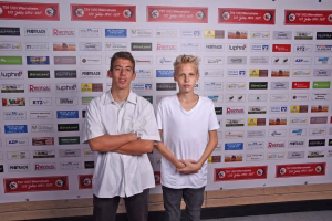 Zwei junge Männer in hellen Shirts posieren nebeneinander vor einer Sponsorwand, Hände ineinander vor dem Körper, im Indoor-Eventbereich.