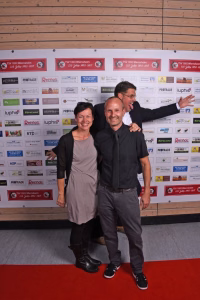 Drei lächelnde Personen posieren vor einer Sponsorenwand bei einer Veranstaltung, eine Person wirkt als Hintergrundfoto-Pose.
