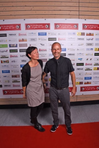 Zwei Personen stehen gemeinsam vor einer Sponsorwand, posieren lächelnd auf rotem Teppich bei einer Veranstaltung.