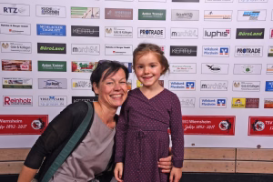Lächelnde Frau mit Kind vor einer Sponsorwand; Arm um das Kind, Fotomoment bei einer Veranstaltung.