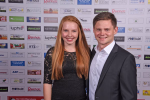 Zwei Personen lächelnd vor einer Wand mit vielen Sponsorlogos; Porträt für Event- oder Pressefoto.