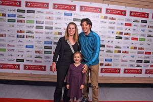 Familie vor Sponsorenwand: zwei Erwachsene und ein Kind lächeln in die Kamera und stehen gemeinsam für ein Erinnerungsfoto.