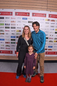 Familie in der Mitte vor einer Sponsorwand; zwei Erwachsene und ein Kind posieren freundlich für ein Erinnerungsfoto bei einer Veranstaltung.