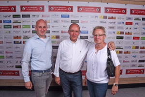 Drei Personen stehen vor einer Sponsorenwand und halten eine Hand am Schulterbereich; Gruppenfoto bei Veranstaltung, mit Bogen in der Bildmitte.