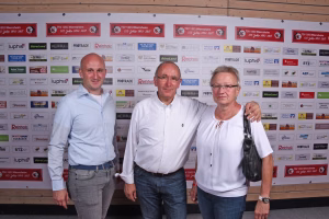 Drei Personen posieren vor einer Sponsorwand mit Logos, in lockerer Kleidung; Blick in die Kamera, Requisit: Handtasche.