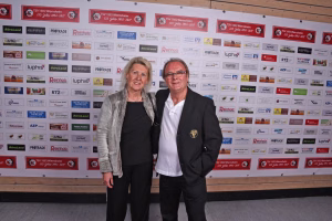 Zwei Personen stehen eng nebeneinander vor einer Sponsorwand mit Logos, lächeln in die Kamera.