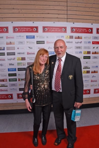 Zwei Personen stehen nebeneinander auf rotem Teppich vor einer Sponsorwand, posieren für ein Event-Foto.