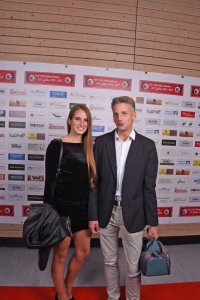 Zwei Personen stehen vor einer Sponsorwand, halten Handtaschen/Umhängetasche und posieren für ein Pressefoto bei einer Veranstaltung.