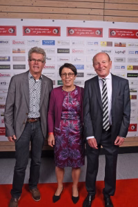 Drei Personen posieren lächelnd nebeneinander vor einer Sponsorwand mit Red-Carpet-Hintergrund.