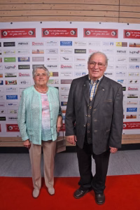 Zwei Personen stehen nebeneinander auf einem roten Teppich vor einer Sponsorenwand und posieren für ein Pressefoto.