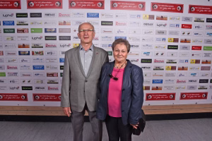 Zwei Personen stehen vor einer Sponsorwand mit Logos; formelle Kleidung, halten Händeschüttel-/Fotopose für ein Eventfoto.