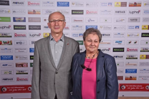 Zwei Personen posieren frontal vor einer Wand mit Sponsorlogos, halten Blick in die Kamera; Hintergrund dient als Veranstaltungs- und Werbefläche.