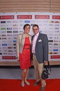Zwei Personen stehen nebeneinander vor einer Sponsorwand mit Logos und Rednerfotowand; sie posieren für ein Eventfoto, eine Person hält eine Tasche.