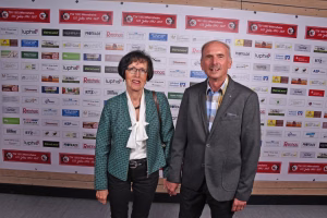 Zwei Personen posieren nebeneinander vor einer Sponsorwand mit Logos; formales Porträt bei Veranstaltung zur Berichterstattung.