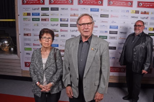 Zwei Personen stehen vor einer bedruckten Fotowand, ein Mann im Anzug und eine Frau im hellen Kostüm, beide schauen in die Kamera.