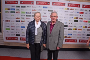Zwei Personen stehen vor einer Sponsorenwand, lächeln in die Kamera; Kleidung für formellen Anlass, Hintergrund dient der Event-Dekoration.