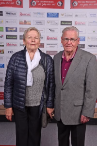 Zwei Personen stehen nebeneinander vor einer Sponsorwand bei einer Veranstaltung, beide blicken in die Kamera und posieren.
