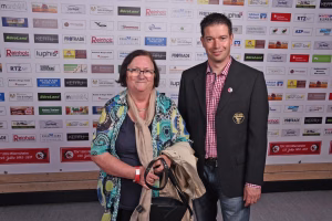 Zwei Personen posieren vor einer Sponsorwand; Person hält ein Stuhlgestell, beide tragen Namensausweis, im Hintergrund Logo-Drucke.