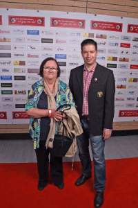 Zwei Personen posieren nebeneinander vor einer Sponsorwand; eine Person hält eine Trophäe, beide lächeln und stehen auf rotem Teppich.
