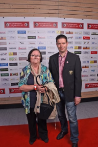 Zwei Personen posieren lächelnd auf rotem Teppich vor Sponsorwand und halten eine Trophäe bzw. Urkunde als Preiserhalt.