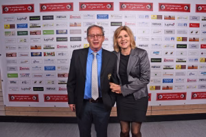 Zwei Personen posieren im Anzug und Kostüm vor einer Sponsorwand mit Logos und Rahmen, für ein offizielles Pressefoto.