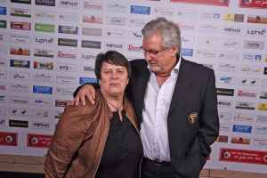Zwei Personen vor einer Sponsorenwand; Mann im Anzug legt den Arm um eine Frau, beide wirken beim Foto posierend.