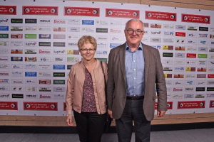 Zwei Personen stehen vor einer Sponsorwand, lächeln in die Kamera; Hintergrund mit vielen Logos und Jubiläumsplaketten.