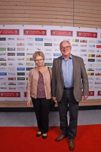 Zwei Personen stehen gemeinsam auf rotem Teppich vor einer Sponsorwand, lächeln und posieren für ein Pressefoto.
