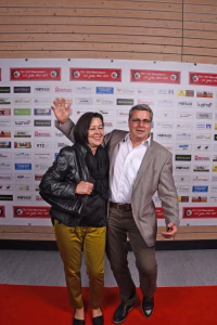 Zwei lächelnde Personen posieren Arm-in-Arm auf rotem Teppich vor einer Sponsorwand als Anlassfoto bei einer Veranstaltung.