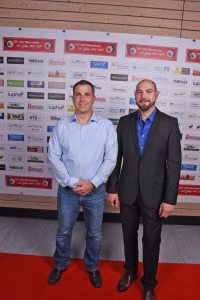 Zwei Männer posieren nebeneinander vor einer Sponsorwand, im Hintergrund Textbanner und Logos als Veranstaltungs-Backdrop.