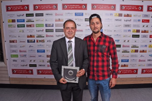 Zwei Personen posieren vor einer Sponsorenwand; eine Person hält eine große Medaille, beide blicken in die Kamera.