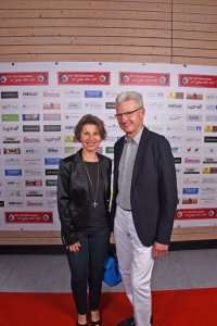 Zwei Personen posieren vor einer Sponsorwand mit Logos; sie stehen auf rotem Teppich und wirken bei einem Empfang/Foto.