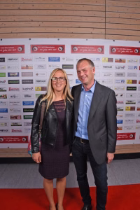 Zwei Personen posieren lächelnd auf rotem Teppich vor einer Sponsorwand, stilgerecht gekleidet; Foto zur Dokumentation eines Events.