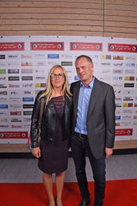 Zwei Personen posieren nebeneinander vor einer Sponsor-Wand; sie stehen lächelnd auf rotem Teppich als offizielles Eventfoto.