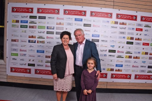 Zwei Erwachsene und ein Kind vor einer Sponsorwand mit Logos, als Gruppenfoto bei einer Feier oder Veranstaltung.