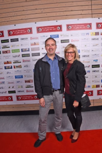 Zwei Personen stehen nebeneinander auf rotem Teppich vor einer Sponsorwand, lächeln und posieren bei einer Veranstaltung.