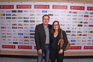 Zwei Personen posieren vor einer Sponsorenwand mit vielen Firmenlogos; eventbezogenes Gruppenfoto bei formellem Anlass.