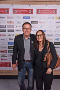 Zwei Personen posieren nebeneinander vor einer Sponsorwand, lächelnd und mit informeller Kleidung; dient als Veranstaltungsfoto.