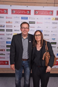 Zwei Personen stehen lächelnd vor einer Sponsorwand mit Firmenlogos; Anlass ist ein gemeinsames Gruppenfoto bei einer Veranstaltung.