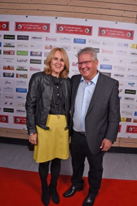 Zwei Personen lächeln nebeneinander auf einem roten Teppich vor einer Sponsorwand, Fotouppe zur Darstellung eines Events.