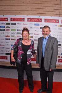 Zwei Personen stehen nebeneinander bei einer Veranstaltung vor einer Sponsorwand, posieren für ein Erinnerungsfoto auf rotem Teppich.