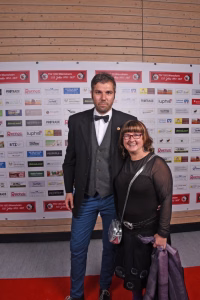 Mann und Frau im festlichen Outfit stehen gemeinsam vor einer Sponsorwand; sie wirken wie bei einer offiziellen Feier, mit Pokal in der Hand.