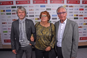 Drei Personen posieren vor einer Sponsorwand bei einer Veranstaltung; im Vordergrund ein Ehepaar und ein weiterer Mann im Anzug.
