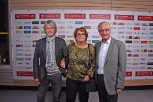 Drei Personen stehen in front eines Sponsor- und Vereinswandbilds; Kleidung und Mimik wirken formell, die Aufnahme dient der Berichterstattung.